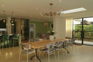 Spacious dining area with a wooden table and decorative chairs, a green-patterned accent wall, pendant lights, and slide-and-turn doors offering a breathtaking view of the garden through large windows.