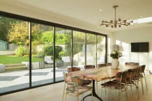 Dining area with a wooden table and floral chairs overlooking a garden through sleek slide and turn doors, illuminated by a modern chandelier above.