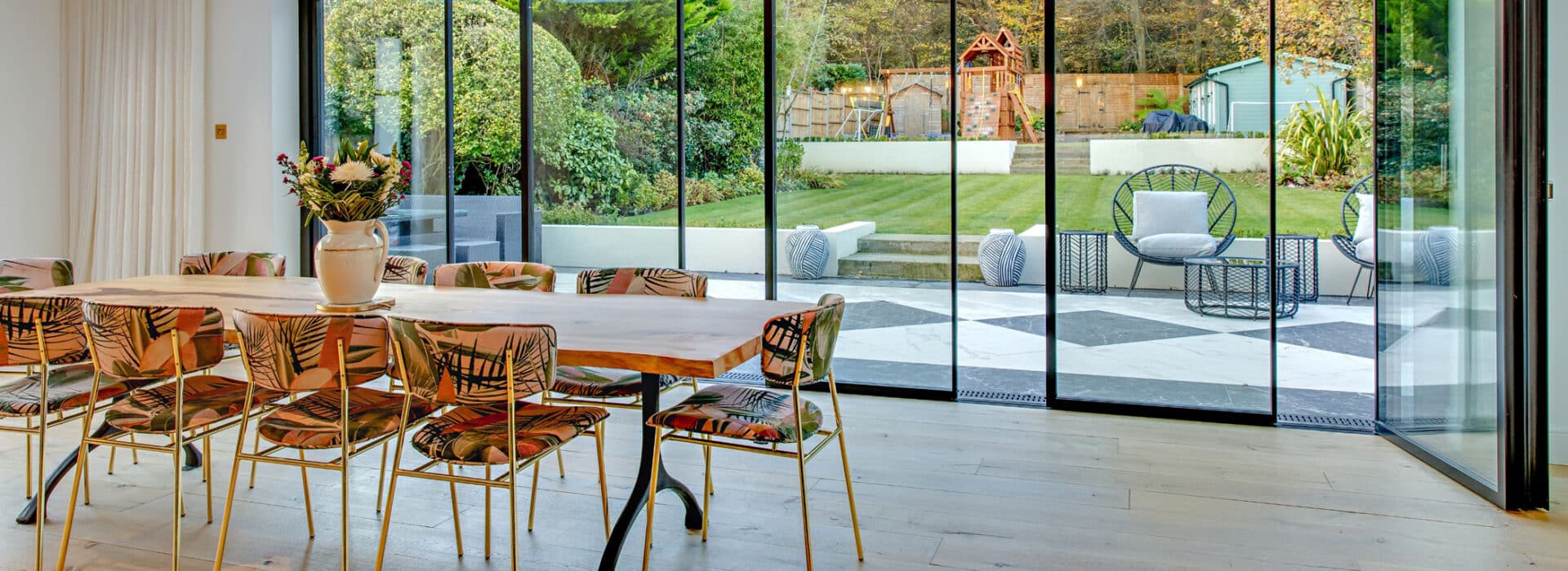 Dining area with a wooden table and decorative chairs in front of sleek slide-and-turn doors leading to a garden with seating, greenery, and a shed.
