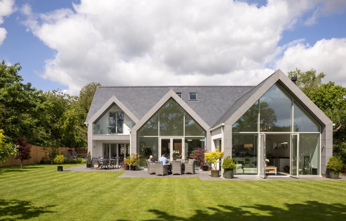 Modern white house with extensive aluminium glazing, bifold doors, and large windows opening onto a manicured garden and patio seating area.