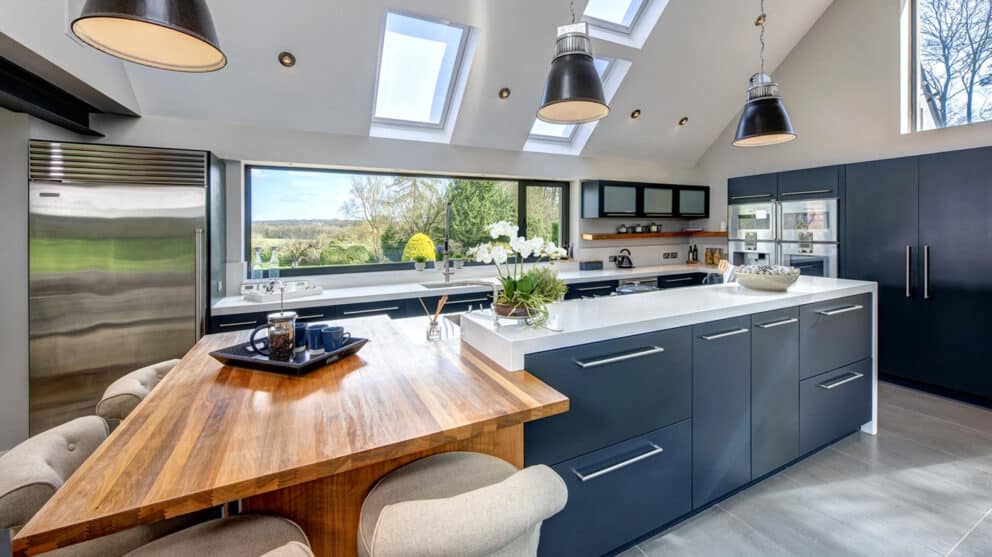 Modern kitchen with a large island, black cabinets, and skylights. Bifold doors open to a wooden dining area, creating a seamless indoor-outdoor flow as natural light streams through the windows, illuminating the space.