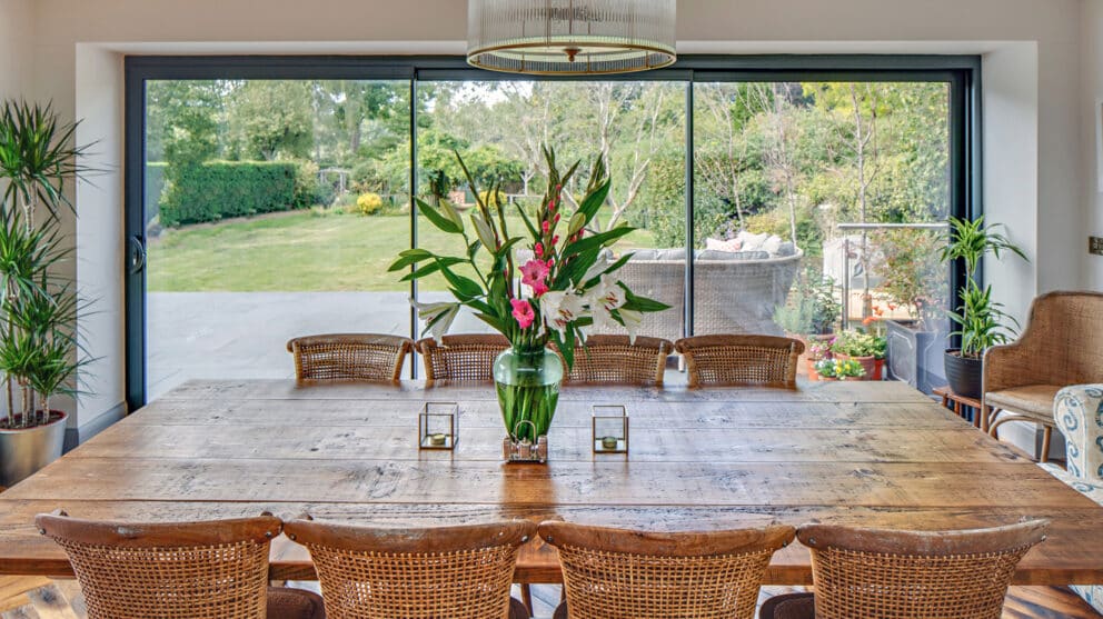 A dining room with a wooden table, wicker chairs, and a vase of flowers features bifold doors that open wide to reveal a garden with a lush lawn and inviting patio furniture.