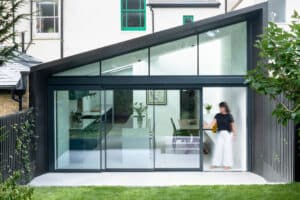 Modern glass extension with a slanted roof, featuring a kitchen and dining area. A person is partially visible near the slimline sliding doors, enhancing the sleek design.