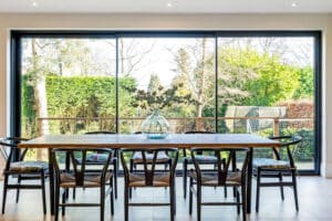 The dining area features a wooden table and six chairs, set before slimline sliding doors that offer an expansive garden view. Centered on the table is a glass vase with elegant branches.