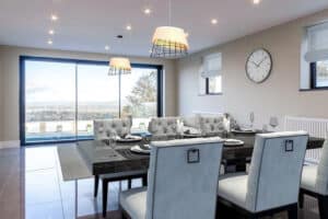 Modern dining room with a large table set for eight. Slimline sliding doors offer a seamless view of the outdoors. Pendant lights hang above, and a clock is on the wall.