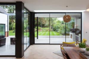 Modern kitchen with slimline sliding doors and glass walls opens to a landscaped garden. There is a wooden dining table, chairs, and an outdoor grill visible under the covered patio.