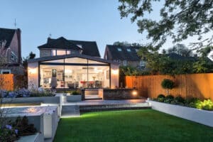 Modern house with large glass windows and slimline sliding doors, illuminated interior, garden with lawn, paved walkway, and wooden fence at dusk.