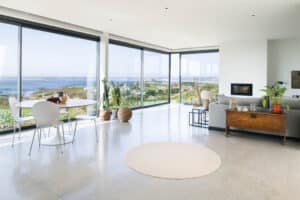 Experience a spacious modern living room with large windows and slimline sliding doors offering a scenic ocean view. The minimalist furniture complements the round dining table and cozy sitting area, creating an inviting and serene atmosphere.