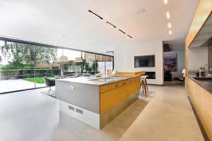 Modern kitchen with a large island, stainless steel appliances, and wooden cabinets. Floor-to-ceiling windows and slimline sliding doors offer a seamless view of the patio and garden.