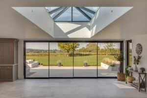 The interior view of the room features slimline sliding doors that open to a lush garden with trees and grass, all beautifully illuminated under a skylight.