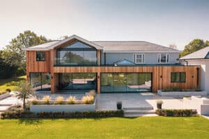 Modern two-story house with large glass windows and slimline sliding doors, a wooden facade on the first floor, surrounded by a well-manicured lawn and patio area.