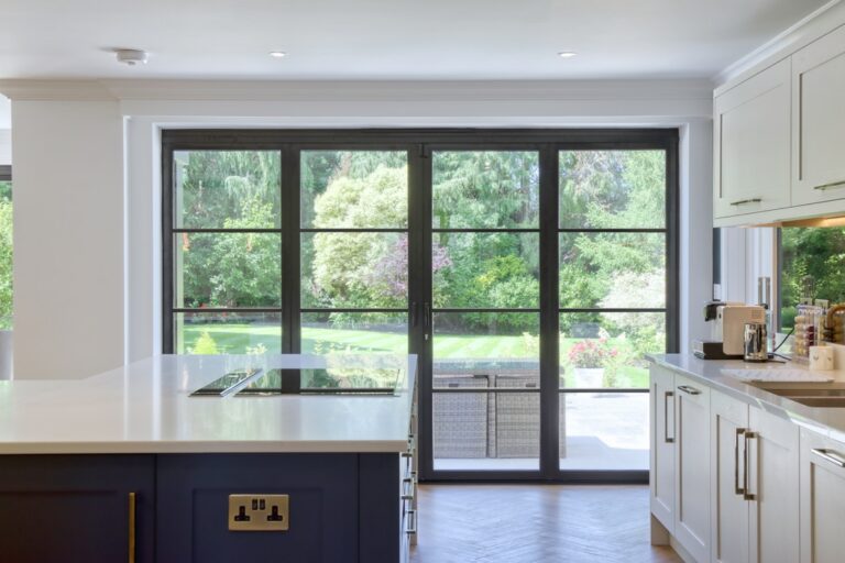 Modern kitchen with a large island and induction cooktop, featuring double glazed bifold doors that open to a garden brimming with trees and shrubs.