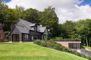 A modern black wooden house with large windows and double glazed bifold doors, surrounded by lush greenery and a neatly maintained lawn. The sky is partly cloudy.
