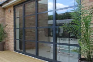 Double glazed bifold doors with a sleek black frame open up to reveal an inviting interior space adorned with bar stools. In the foreground, you'll find potted plants and a wooden deck, creating a seamless blend of indoor and outdoor living.