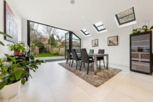 Modern dining room with table and six chairs on a rug, framed by double glazed bifold doors that open to a garden. A wine cooler sits discreetly on the right, while natural light from skylights enhances the bright, inviting space.