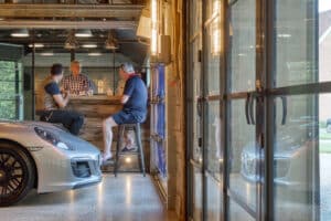 Three men sit at a bar inside a garage, with double glazed bifold doors offering a clear view of a silver sports car parked nearby.