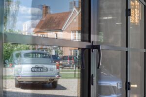 Reflection of a vintage silver car in double glazed bifold doors, with a brick house and greenery visible in the background.