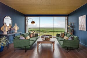 Modern living room with green sofas, wooden coffee table, large windows with slimline sliding doors showcasing a scenic view, blue walls, and decorative elements including lamps, plants, and wall art.