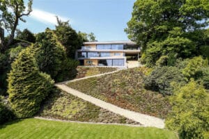 A modern multi-story house with large windows and sleek slimline sliding doors, surrounded by a well-maintained garden and lush trees on a sloped terrain. A pathway gracefully leads up to the entrance.