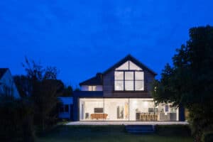 Modern house with a glass facade and sleek slimline sliding doors, illuminated at night. It showcases a spacious interior featuring a pool table, dining area, and kitchen. Surrounded by trees under a dark blue sky.