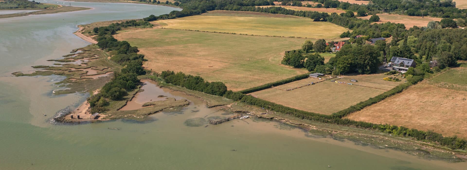 An aerial view of a rural landscape reveals lush green fields, scattered houses, and a narrow river coastline with sandy and rocky areas. Through the reflections in 4 panel sliding glass doors, this serene vista whispers tales of tranquility and untamed beauty.