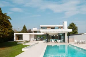 Modern house with large windows, flat roof, and a swimming pool in a landscaped garden with trees and outdoor seating.