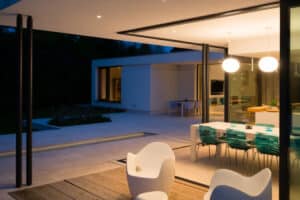 Modern open-plan house with glass walls, white furniture, and patio. Indoor dining area with green seats and round lights. Evening lighting creates a cozy ambiance.