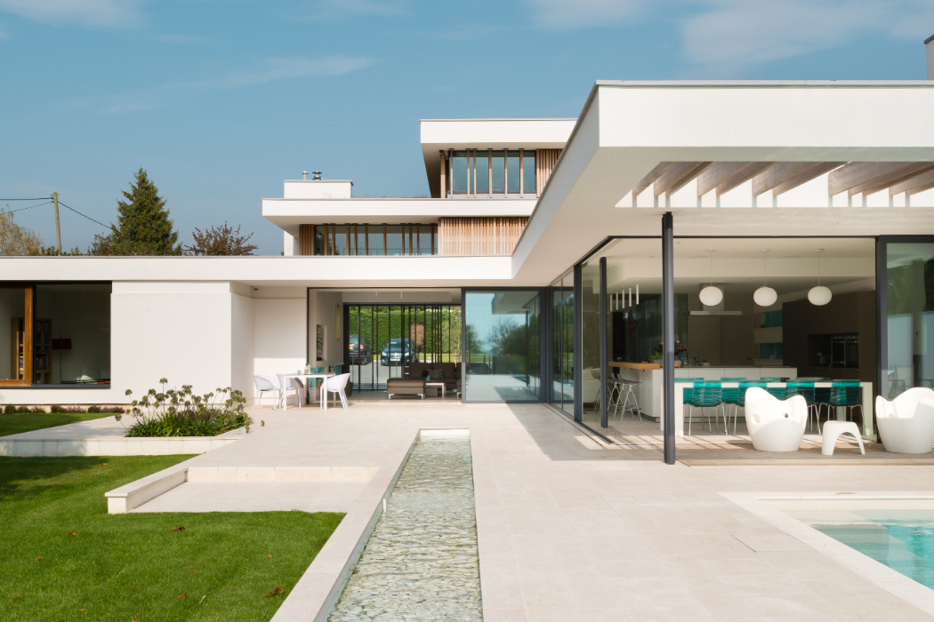 Modern white house with large glass windows, a sleek patio, outdoor seating, and a pool, set against a clear blue sky.