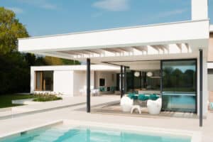 Modern house with a large overhanging roof, glass walls, and outdoor seating by a pool. Minimalist design with white walls and turquoise accents.