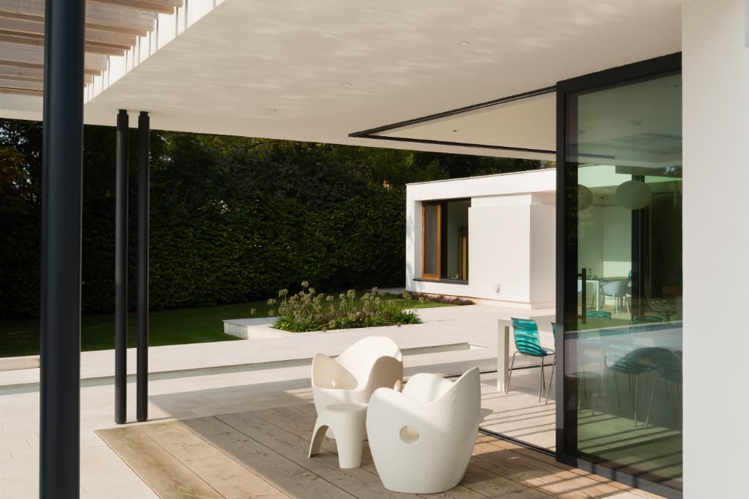Modern patio with white chairs, wooden decking, and a garden view. Large glass doors open to reveal an interior space with a blue chair and table.