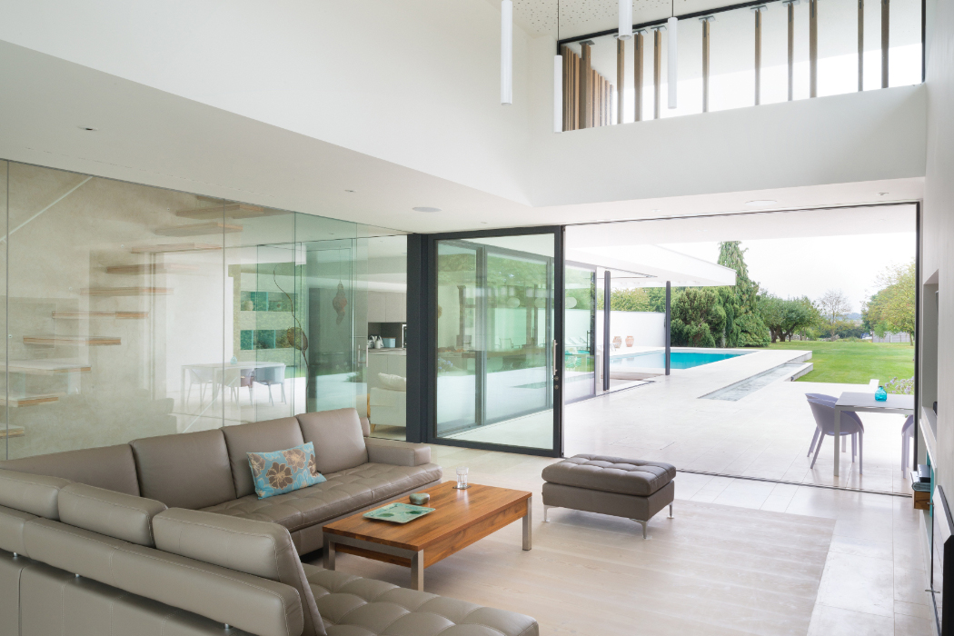Modern living room with beige sectional sofa and glass walls opening to a patio with a pool and garden view. A wooden coffee table and a small ottoman are in the seating area.
