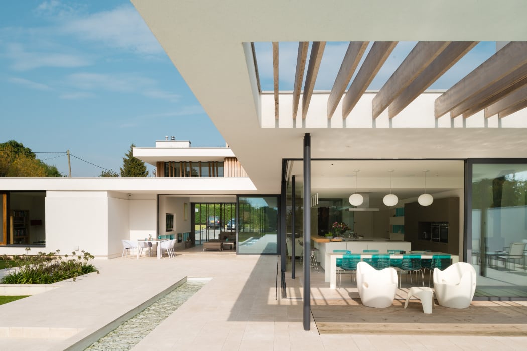 Modern house with open patio, glass walls, and minimalist furniture. Patio features seating area and dining set. Pool seen along the tiled ground. Clear sky background.