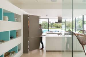 Person walking through a pivot door in a modern kitchen with white cabinets, open shelving, and a view of a backyard pool through large glass windows.
