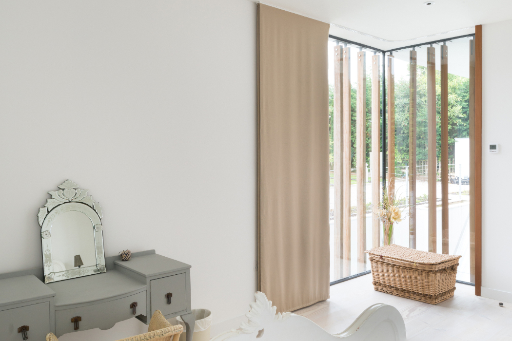 A corner of a bright room with a small grey vanity, mirror, cushioned stool, wicker basket, and large sliding glass doors with vertical wooden slats. A view of greenery outside.