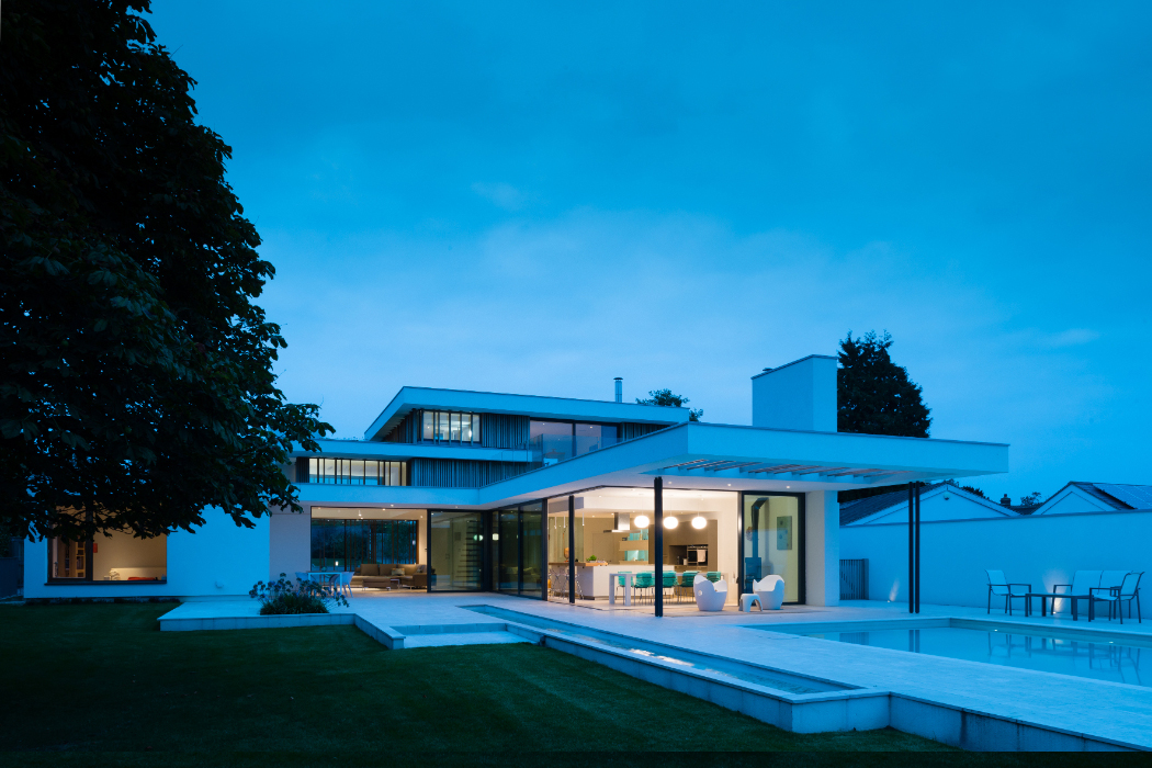 Modern house with large glass windows and a flat roof, surrounded by a lawn. A pool reflects the exterior lighting in the foreground, under a dusky sky.