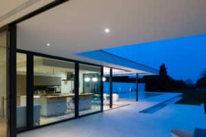 Modern house with large glass windows showing kitchen and dining area, adjacent to a pool and patio, during twilight.