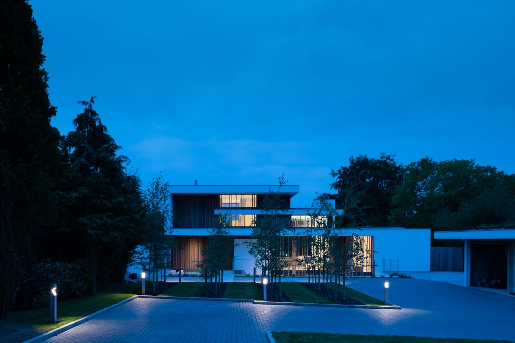 Modern house with illuminated windows at dusk, surrounded by trees and a paved driveway, under a blue evening sky.