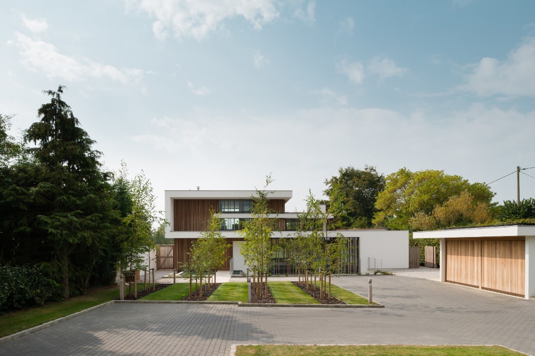 Modern two-story house with large windows, surrounded by trees and a paved driveway. A separate garage is on the right, with clear skies above.