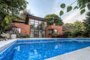 Modern house with large windows, surrounded by trees, featuring a rectangular swimming pool in the foreground.