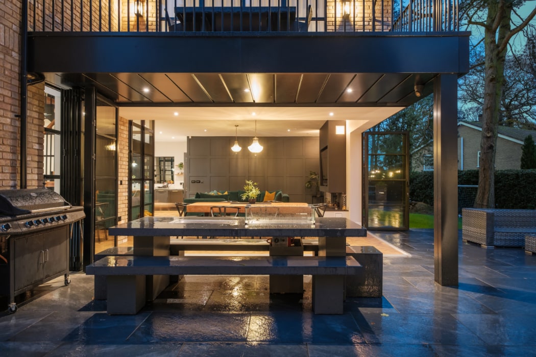 Modern outdoor dining area with a large table and grill, adjacent to a well-lit indoor living space with glass doors on a rainy evening.