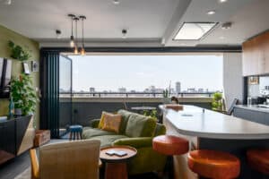 Modern open-plan living room with green sofa, kitchen island, and bar stools. Large folding doors open to a balcony with city views. Indoor plants and ceiling lights complement the decor.