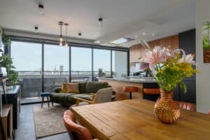 Modern open-plan living space with a wooden dining table, green sofa, and large windows providing a city view. A vase with flowers is on the table, and kitchen area is in the background.