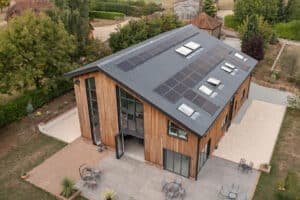 Aerial view of a modern house with a slanted roof, covered in solar panels. The building features curtain walling, wooden siding, large windows, and a spacious patio with outdoor furniture.