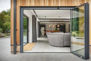 Open glass doors reveal a modern living room with a curved gray sofa, pendant lights, and a dining area. The patio outside leads to a lush green garden.