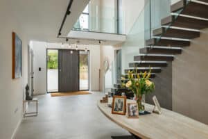 Modern foyer with minimalist decor, glass staircase, and photo-laden round table. Large front door with sidelights opens to a bright space, artwork on walls, and flowers on the table.