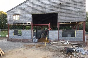 A wooden barn under construction with brick walls and curtain walling being installed. Scaffolding and construction materials are visible around the site.