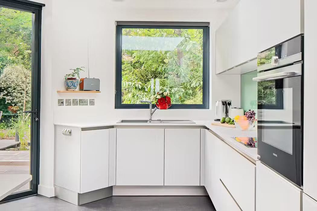 Modern white kitchen featuring sleek aluminium casement windows, overlooking greenery. A potted plant and a red vase adorn the counter, with an oven and cabinets visible. The door to the left opens to a garden.