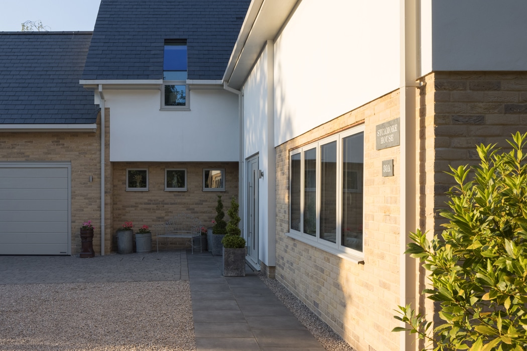 Modern two-story house with a brick and white exterior, large aluminium casement windows, a garage, and potted plants along the paved entrance.