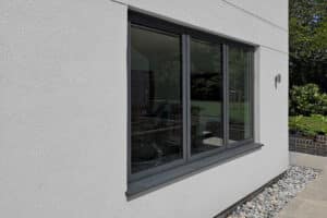 An aluminium casement window with dark gray frames sits on a white stucco wall, overlooking a garden with trees and grass. Pebbles line the base of the wall.