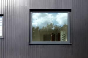 An aluminium casement window on a dark gray paneled wall reflects a cloudy sky and trees.